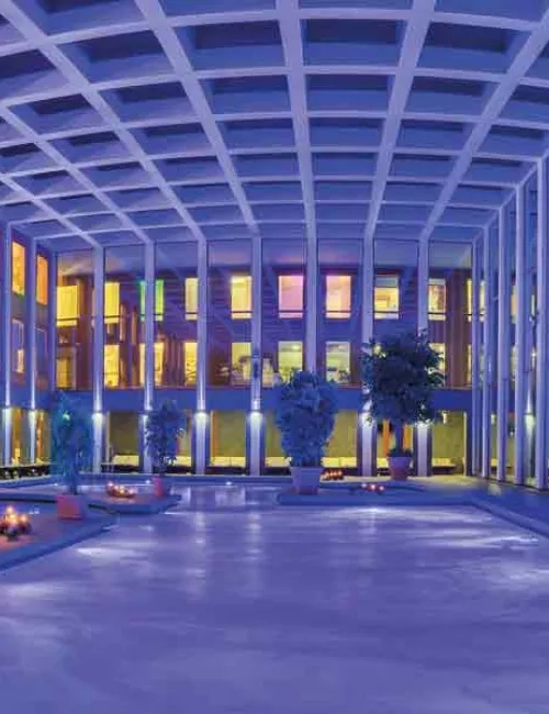 Innenbereich der SaarowTherme mit beleuchtetet Becken Abends. Dekorative Elemente wie Grünpflanzen in verschiednenen Größen und angezündete Kerzen am Beckenrand platziert. Das Solewasser ist durch die Unterwasserscheinwerfer lila gefärbt und daher wirkt das Bild blau lila. Helles bis goldenes Licht scheint durch die Glasfront vom Eingangsbereich im oberen teil des Gebäudes.