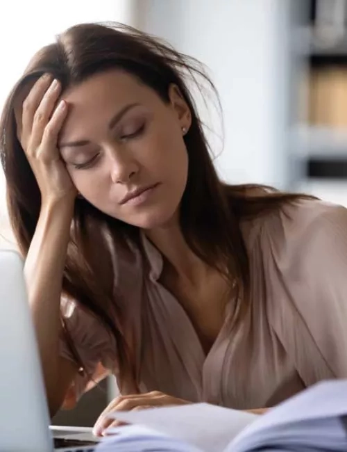 Eine Frau mit braunen, langen Haaren, sitzt an einem vollen Schreibtisch und stützt sich erschöpft den Kopf auf der Hand ab. Dabei hat sie ihre Augen geschlossen.