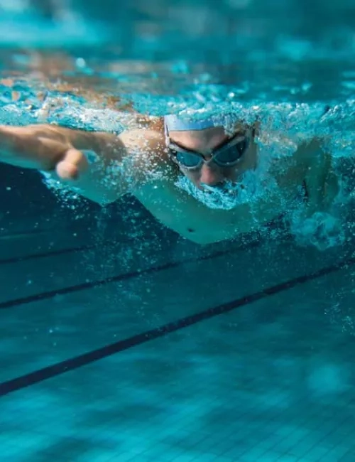Ein Leistungsschwimmer mit Schwimmbrille und Badekappe wird unter Wasser in einem Sportbecken abgebildet. Er krault und hat seinen rechten Arm vorne. 