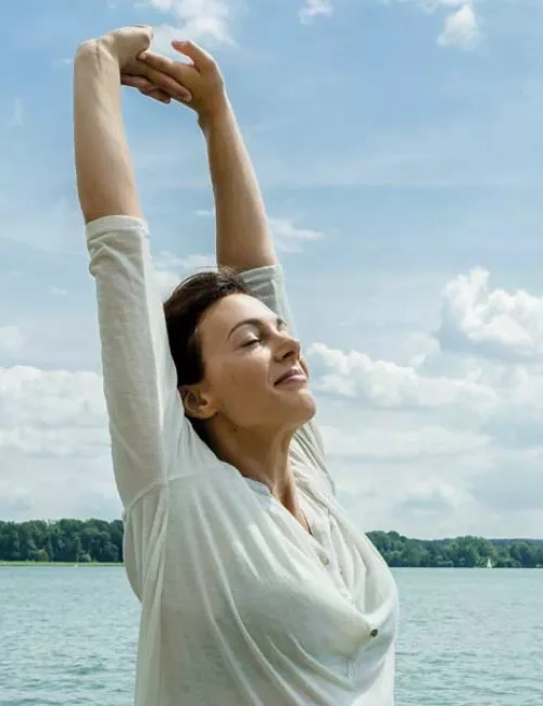 Eine Frau mit dunklen Haaren und einem weißen langärmligen Shirt steht am See, streckt die Arme in den Himmel und ihr Gesicht zeigt in Richtung Sonne mit geschlossenen Augen. Ein leichtes lächeln ist zu erkennen. Im Hintergrund sind man Wald und davor den See mit Segelschiffen.