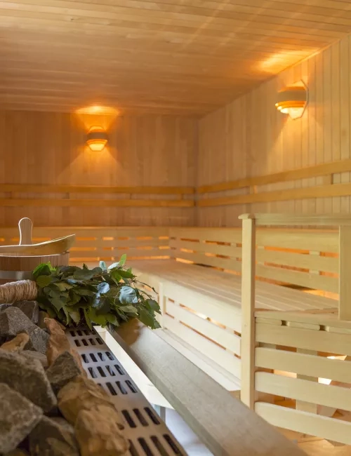 Helle, moderne Hiki Sauna mit Holzverkleidung und mehreren Sitz- bzw. Liegeebenen; im Vordergrund ein Saunaofen mit Lavasteinen und einem Aufgusskübel.