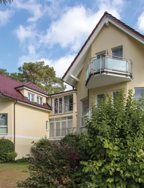 Zweistöckiges hellgelbes Wohnhaus mit roten Dächern und kleinen Balkonen, umgeben von Grünpflanzen und Bäumen unter blauem Himmel.