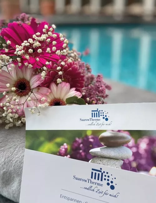 Gutscheine der SaarowTherme an einem weißen Handtuch drapiert. Im Hintergrund ist das Thermalbad der SaarowTherme mit türkisblauem Wasser zu sehen. Blumen am Handtuch gelegt deuten auf Geschenkgutscheine hin.