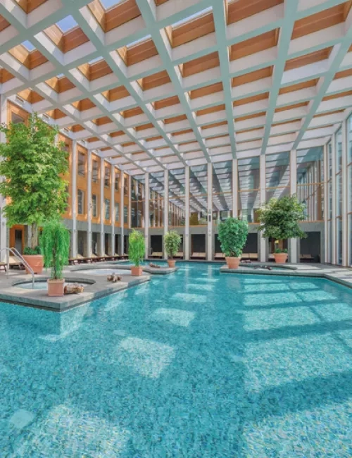 Lichtdurchflutetes Thermalbad mit großen Fenstern und Deckenkuppel. Türkisblaues Wasser und grüne Pflanzen vermitteln ein Ort zum Entspannen und Erholen.