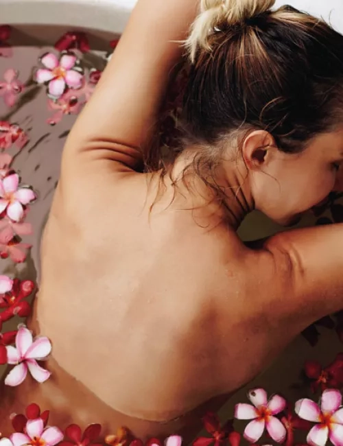 Eine Frau entspannt in einer runden Badewanne mit warmem Wasser und schwimmenden Blüten. Sie lehnt sich mit geschlossenen Augen und erhobenen Armen an den Wannenrand, umgeben von Wellnessprodukten. Die Atmosphäre wirkt ruhig und erholsam.