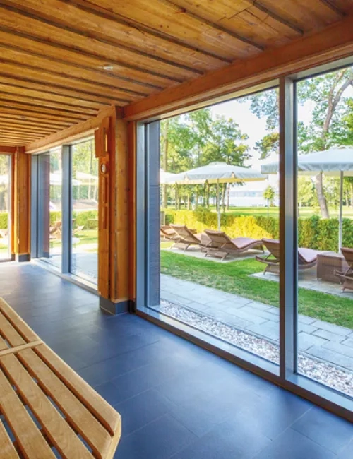 Innenansicht der großzügigen, lichtdurchfluteten KeloSauna mit Holzverkleidung und Panoramafenster. Draußen sind Liegestühle und Sonnenschirme im gepflegten Gartenbereich zu sehen.