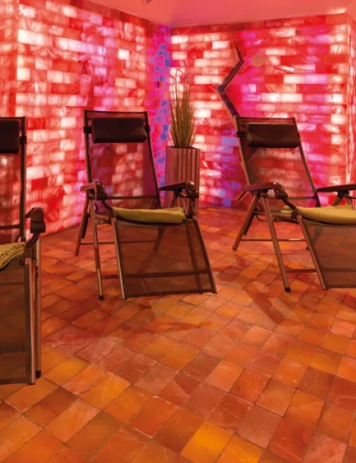 Mehrere Relaxliegen stehen in einem stimmungsvoll beleuchteten Ruheraum mit roten Salzwänden und warmem Licht. Der Boden ist mit Salzsteinen ausgelegt, an den Wänden stehen dekorative Pflanzen.