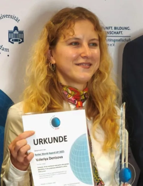 Junge Frau mit lockigen roten Haaren hält eine Urkunde in den Händen und lächelt in die Kamera, flankiert von zwei Männern im Anzug bei einer offiziellen Preisverleihung vor einer Logo-Fotowand.