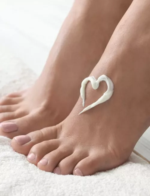 Zwei gepflegte, barfuß stehende Füße auf einem weichen weißen Handtuch. Auf dem rechten Fuß ist ein Herz aus Creme aufgetragen. Die Szene vermittelt Ruhe und Selbstfürsorge.
