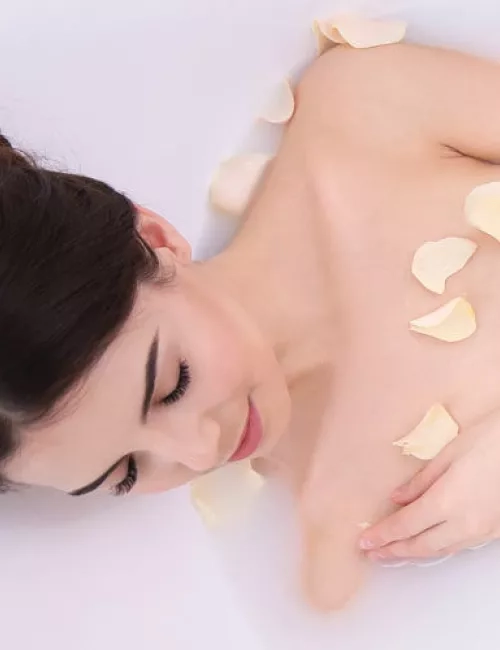 Eine Frau mit dunklen Haaren entspannt in einem Milchbad mit hellen Rosenblättern auf der Haut.