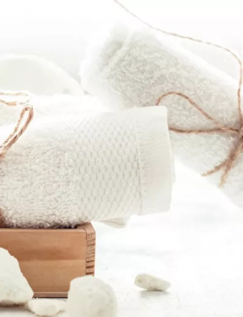 Weißes Handtuchset mit dekorativer Kordel auf einem Holztablett, daneben weiße Naturseife und Bimssteine.