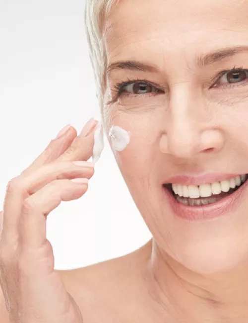 Eine lächelnde ältere Frau mit kurzen grauen Haaren trägt sich sanft Gesichtscreme auf die Wange auf. Sie blickt direkt in die Kamera und vermittelt ein Gefühl von Zufriedenheit und Pflege.