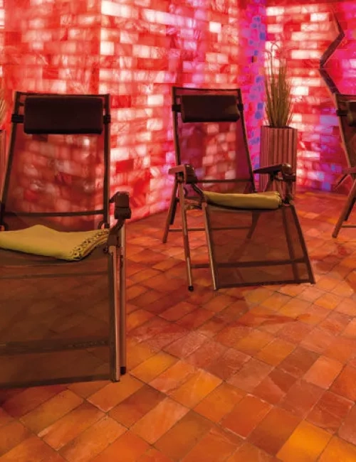 Mehrere Relaxliegen stehen in einem stimmungsvoll beleuchteten Ruheraum mit roten Salzwänden und warmem Licht. Der Boden ist mit Salzsteinen ausgelegt, an den Wänden stehen dekorative Pflanzen.