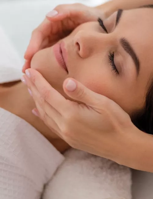 Eine entspannte Frau mit geschlossen Augen erhält eine sanfte Gesichtsmassage in wohltuender Umgebung.