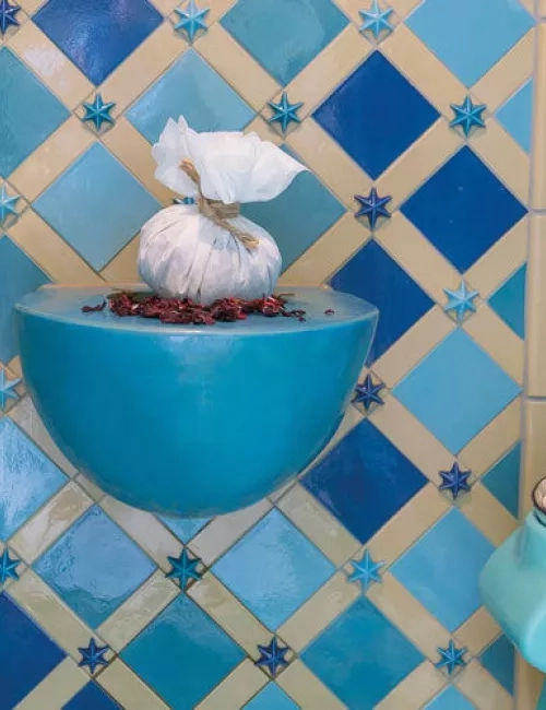 Blick auf einen stilvoll gestalteten orientalischen Ruhebereich mit türkisfarbenen Liegeflächen und einer kunstvoll gefliesten Wand in Blau- und Beigetönen. In der Mitte befindet sich ein dekorativer Wasserbrunnen.