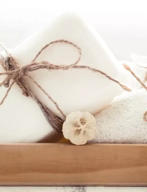 Ein dekoratives Arrangement aus weißer Seife, gebunden mit rustikalem Juteband, daneben ein weiches, gefaltetes Handtuch und eine kleine getrocknete Blume. Alles präsentiert auf einem Holztablett in heller, natürlicher Umgebung.