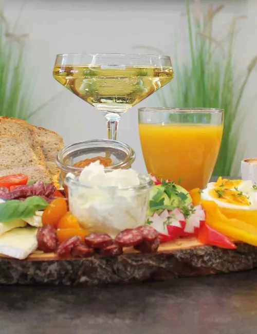 Ein liebevoll angerichtetes Frühstücksbrett mit Aufschnitt, Käse, frischem Gemüse, Obst und Dips steht auf einem rustikalen Holztablett. Daneben ein Glas Orangensaft, ein Glas Sekt und ein Korb mit Körnerbrot. Im Hintergrund ein stilvoll eingerichteter Raum mit Holzverkleidung und dekorativen Schneidebrettern an der Wand.