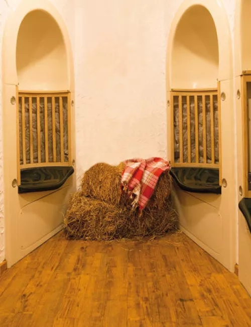 Ein gemütlich eingerichteter Ruheraum mit Holzfußboden, halbrunden Nischen mit gepolsterten Sitzflächen und Heuballen, auf denen eine rot-karierte Decke liegt. Die Atmosphäre ist warm und einladend.