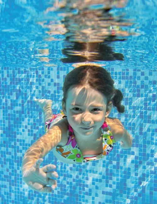 Ein Mädchen in buntem Badeanzug schwimmt unter Wasser in einem Pool mit blauen Mosaikfliesen und schaut lächelnd zur Kamera.