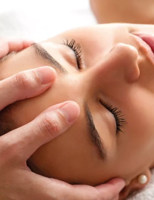 Das Bild zeigt eine Nahaufnahme einer Person, die eine Gesichtsmassage erhält. Die Augen sind geschlossen, während eine andere Person mit den Händen sanft das Gesicht massiert. Die Szene wirkt entspannend und erinnert an eine Spa- oder Kosmetikbehandlung mit ruhiger, beruhigender Atmosphäre.