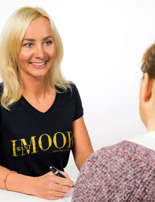 Zwei Frauen im Gespräch, eine davon trägt ein schwarzes T-Shirt mit der Aufschrift „I WANT MOOR“ und hält einen Stift in der Hand.