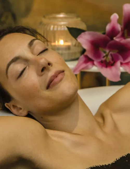 Frau entspannt mit geschlossenen Augen in einer Badewanne, Kerze und Blume im Hintergrund.