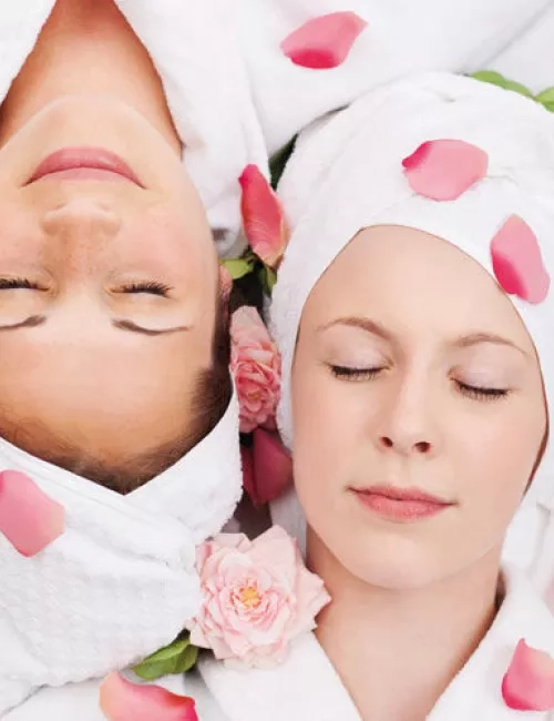 Zwei Frauen in weißen Bademänteln und Handtuchturbanen liegen entspannt mit geschlossenen Augen, umgeben von Rosenblättern und Blüten.