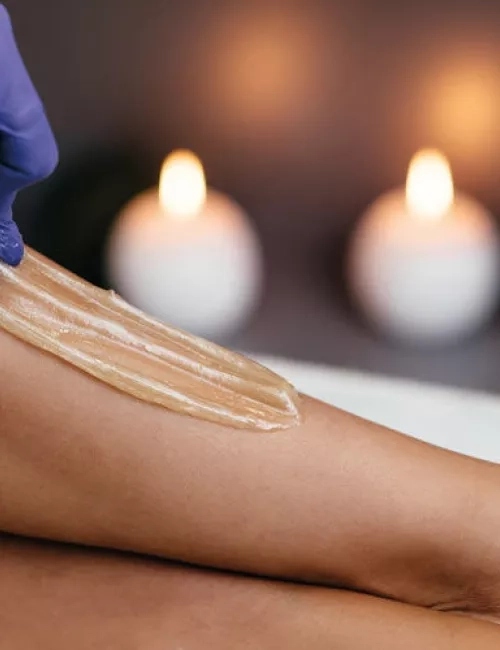 Auftragen von Zuckerpaste auf ein Bein zur Haarentfernung in entspannter Atmosphäre mit Kerzen im Hintergrund.