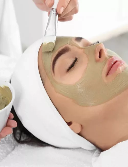 Eine Frau liegt entspannt auf einer Liege mit geschlossen Augen, während ihr mit einem Pinsel eine grüne Gesichtsmaske aufgetragen wird. Sie trägt ein weißes Haarband und ist in ein weißes Handtuch gehüllt.