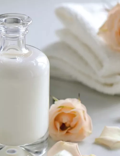 Eine elegante Glasflasche mit weißer Lotion steht auf einem hellen Untergrund. Umgeben ist sie von zarten Rosenblüten und weißen Handtüchern, was eine ruhige, pflegende Spa-Atmosphäre vermittelt.