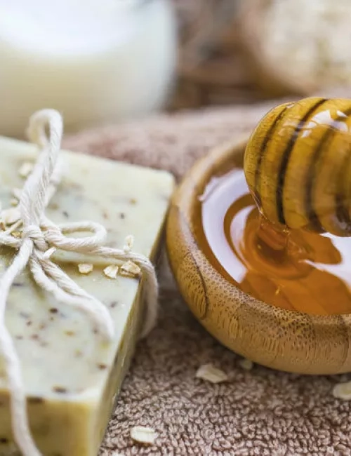 Ein Stück handgemachte Seife mit Haferflocken liegt auf einem Handtuch neben einer Holzschale mit flüssigem Honig und einem Honiglöffel.