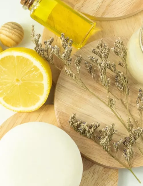 Arrangement aus Naturkosmetik-Zutaten: eine aufgeschnittene Zitrone, Massagekugeln aus Holz, ein Glas mit gelbem Öl, getrocknete Lavendelzweige und weitere Naturprodukte auf runden Holzunterlagen.