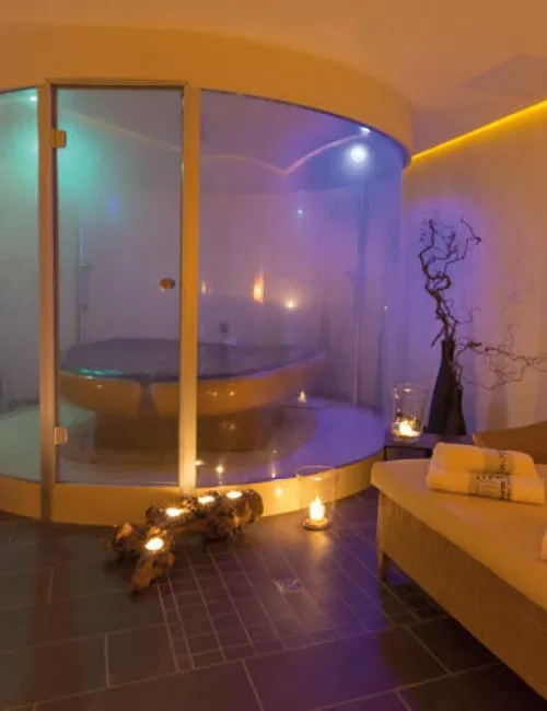 Ein stilvoll beleuchteter Wellnessraum mit einer modernen Membranliege in einer gläsernen Kabine. Umgeben von dekorativen Elementen wie Ästen und Kerzenlicht entsteht eine warme und entspannte Atmosphäre. Daneben liegt ein Handtuch auf einer bequemen Duoliege.