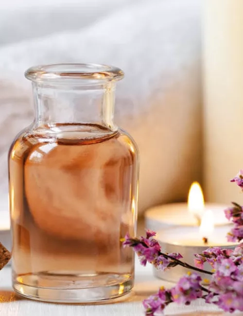 Ein Glasfläschchen mit rosafarbenem Öl steht auf einem Tisch, umgeben von brennenden Teelichtern, einem Handtuch, einem Korken und violetten Blüten.