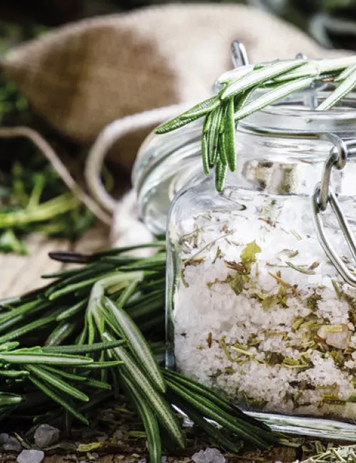 Ein Glas mit grobem Meersalz und getrockneten Kräutern, verziert mit frischem Rosmarin. Im Hintergrund liegen weitere Kräuter und ein Jutesäckchen, was eine natürliche und aromatische Atmosphäre vermittelt.