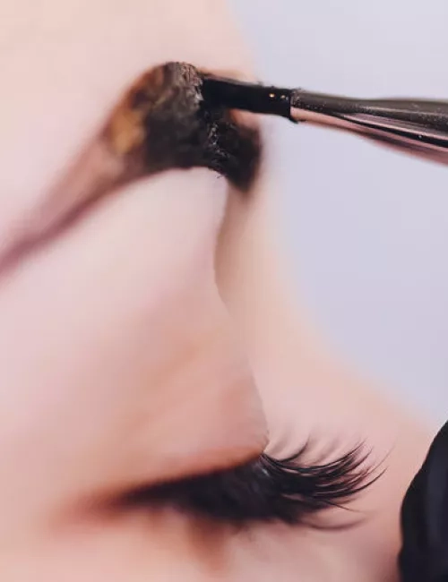 Nahaufnahme eines geschlossenen Auges mit langen Wimpern, während mit einem Pinsel dunkle Farbe auf die Augenbraue aufgetragen wird.