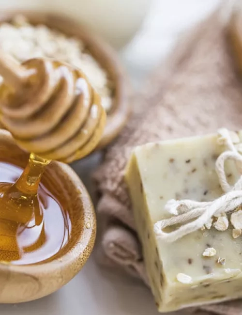 Naturseife mit Haferflocken dekoriert, daneben eine Schale mit flüssigem Honig und ein Honiglöffel arrangiert auf einem weichen Handtuch.