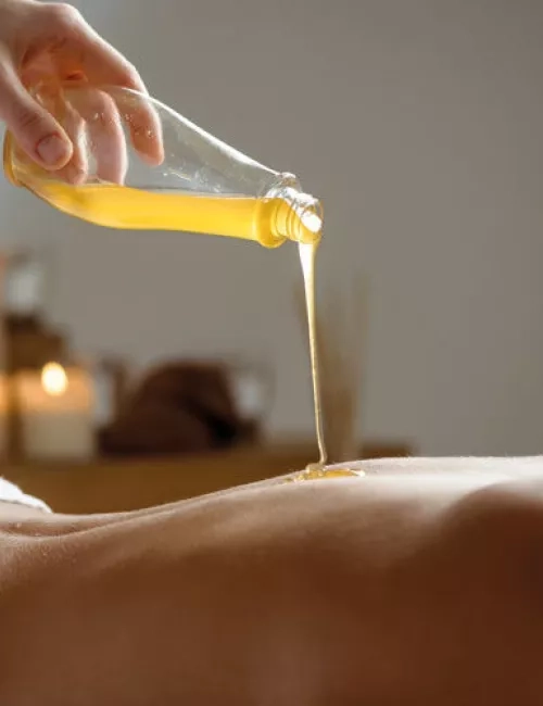Eine Hand gießt warmes Massageöl aus einer Glasflasche auf den Rücken einer liegenden Person. Im Hintergrund sorgen brennende Kerzen für eine entspannte und wohltuende Atmosphäre.