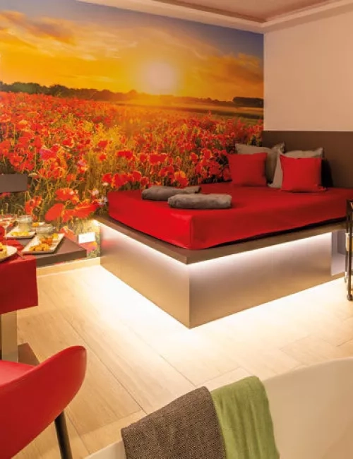 Ausschnitt aus dem Privat Spa. Ein Wasserbett mit roten Dekokissen und einer grauen Kuscheldecke. Das Bett wird von unten mit einem Lichtschlauch warm gelb beleuchtet. Auf der Hintergrundwand ist ein Sonnenuntergang zu sehen und ein Kornblumenfeld in Rot abgebildet. Zwei rote Sesselartige Stühle mit einem gedeckten Tisch.