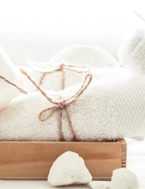 Weißes Handtuchset mit dekorativer Kordel auf einem Holztablett, daneben weiße Naturseife und Bimssteine.