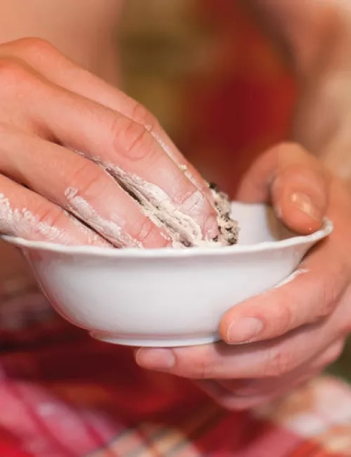 Ein Mann trägt in einem Wellnessbereich mit rotem Saunatuch Heilschlamm aus einer kleinen weißen Schale auf seinen Arm auf. Im Hintergrund ist unscharf eine weitere Person mit ähnlichem Tuch zu sehen, die ebenfalls Schlamm aufträgt.