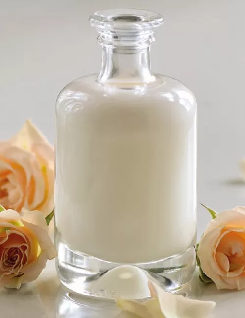 Eine elegante Glasflasche mit weißer Lotion steht auf einem hellen Untergrund, umgeben von zartrosa Rosen und Rosenblättern.