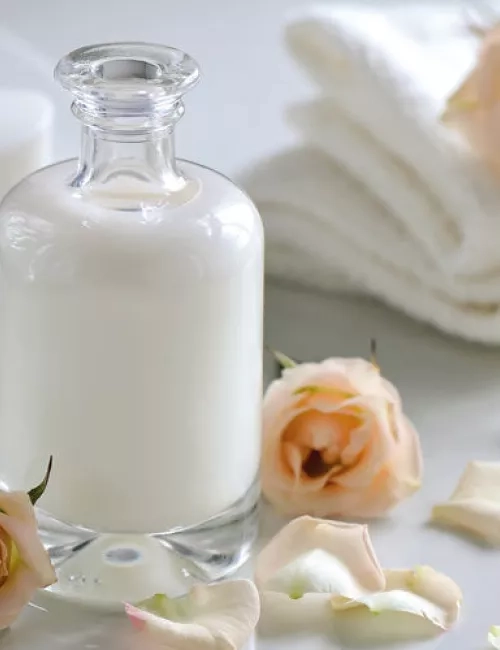 Eine elegante Glasflasche mit weißer Lotion steht auf einem hellen Untergrund. Umgeben ist sie von zarten Rosenblüten und weißen Handtüchern, was eine ruhige, pflegende Spa-Atmosphäre vermittelt.