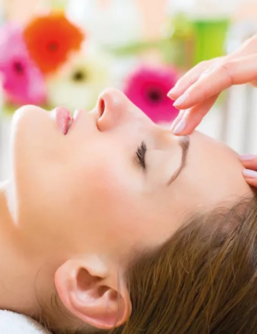 Eine Frau liegt entspannt mit geschlossenen Augen auf einer Wellnessliege, während eine Hand sanft ihre Stirn massiert. Im Hintergrund sorgen bunte Blumen für eine ruhige und angenehme Atmosphäre.