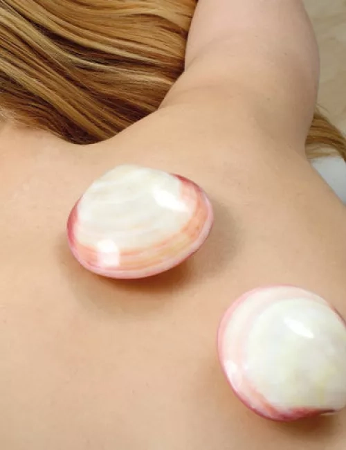 Eine Frau liegt mit dem Rücken nach oben auf einer Wellnessliege. Auf ihrem Rücken befinden sich zwei große, glänzende Muschelschalen. Die Muscheln sind rosa-weiß gemustert und wirken wohltuend auf der Haut.