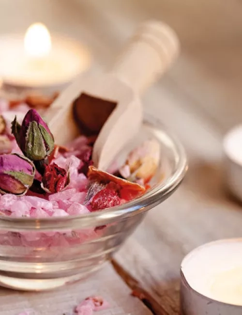 Eine Glasschale mit rosa Badesalz, Rosenknospen und einer kleinen Holzschaufel, umgeben von brennenden Teelichtern auf einem Holztisch.