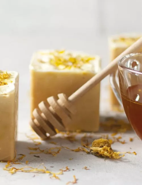 Handgemachte Seifenstücke mit getrockneten Blüten, daneben eine Tasse mit Honig und ein Holz-Honiglöffel.