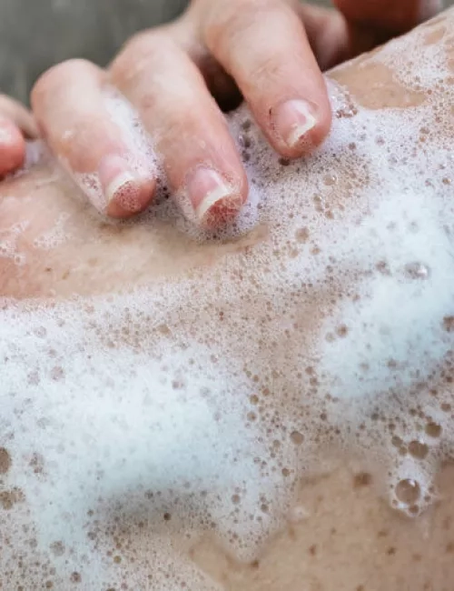 Eine Hand wäscht einen mit Schaum bedeckten Rücken. Die Nahaufnahme zeigt dichte Seifenblasen auf der Haut, die eine Reinigung oder Massage andeuten.