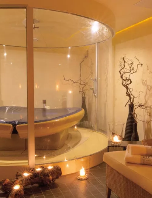 Ein stilvoll beleuchteter Wellnessraum mit einer modernen Wasserliege in einer gläsernen Kabine. Umgeben von dekorativen Elementen wie Ästen und Kerzenlicht entsteht eine warme und entspannte Atmosphäre. Daneben liegt ein Handtuch auf einer bequemen Liege.