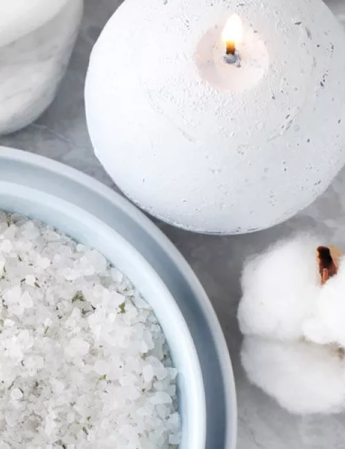 Eine weiße Kugelkerze brennt auf einem hellen Untergrund. Daneben stehen eine Schale mit grobem Badesalz und eine weiche Baumwollblüte.
